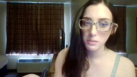 Media: Video of a young woman with fair skin, long brown hair, and rectangular glasses, wearing a light blue tank top, standing in a room with brown patterned curtains and a wooden floor.