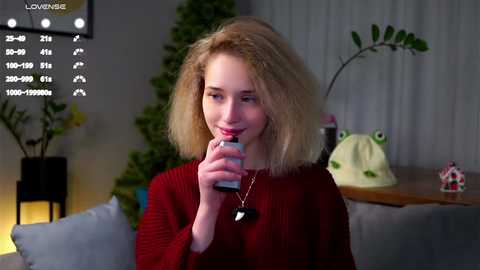 Video of a young woman with blonde hair, fair skin, wearing a red sweater, drinking from a can in a cozy, dimly lit living room with green plants and a frog toy.