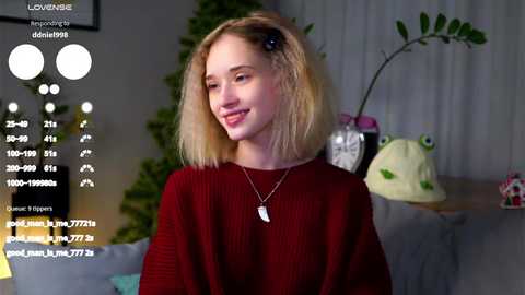 Video of a young woman with shoulder-length blonde hair, wearing a maroon sweater, standing indoors beside a green plant and a lamp. The background includes a white curtain.