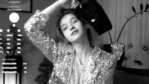 A black-and-white video of a young woman with short, tousled hair, wearing a textured, sequined jacket, and a fur hat, posing with a relaxed expression, surrounded by studio lights and a large plant.