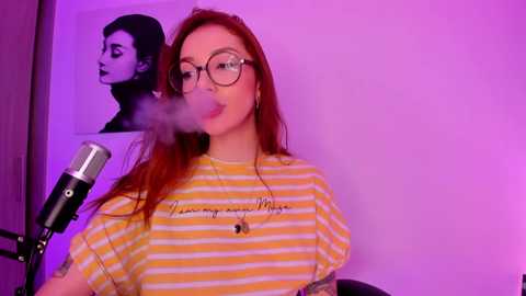 Video of a red-haired woman in glasses, blowing smoke rings, wearing a yellow-striped T-shirt with a tattooed arm, against a purple-lit background with a black-and-white Marilyn Monroe poster.