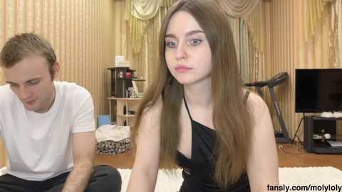 Media: Video of a young woman with long brown hair, wearing a black dress, and a young man with short blond hair, both sitting on a rug in a cozy, wooden-paneled room.