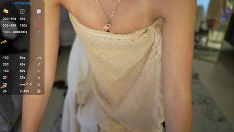 Video of a slender, fair-skinned woman wearing a beige, patterned, strapless dress, featuring a dainty necklace, in a blurred indoor setting with a virtual camera interface on the left.