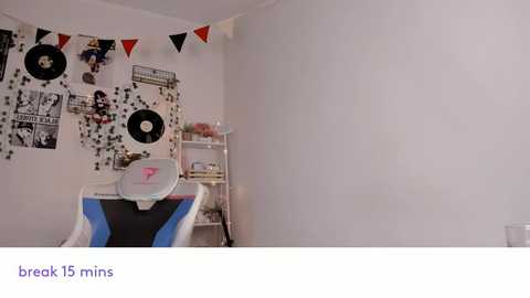 Video of a minimalist, white-walled room with a gaming chair, white shelving unit, and a collage of black-and-white vinyl records and posters.