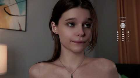 Media: A video of a young woman with fair skin and dark brown hair, wearing a necklace, in a dimly-lit room with modern abstract art on the wall and beige curtains.