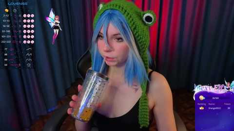 A video of a woman with blue hair, wearing a green frog hat, drinking from a glass, in a dimly lit room with red curtains and a black chair.