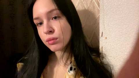 Media: A video of a young woman with long black hair, fair skin, and a neutral expression, wearing a floral-patterned top, standing in a dimly lit room with textured walls.