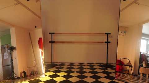 Media: Video of a small, sparsely furnished room with a checkered black and white floor. It features a pair of wooden ballet bars, a red folding chair, and a cluttered corner with miscellaneous items.