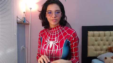 Media: A video of a woman with long, dark hair wearing a Spider-Man suit and glasses, standing in a modern bedroom with a tufted headboard and a window with blinds.