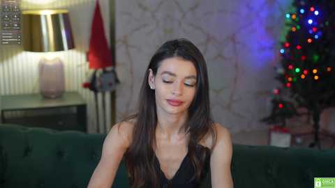 Media: A video of a young woman with long, straight black hair, wearing a black dress, sitting on a green couch, in a room with a decorated Christmas tree, a lit lamp, and patterned wallpaper.