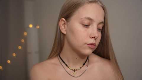 Media: Video of a young Caucasian woman with straight blonde hair, fair skin, and minimal makeup. She wears a black choker necklace and a silver chain necklace with a pendant, looking introspective. Background is blurred with warm, ambient lighting.