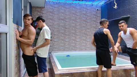 Video of four shirtless men in black shorts standing at an indoor pool with a brick wall background. One man touches a wall, another holds a phone, another looks at the pool, and the fourth leans against a railing.