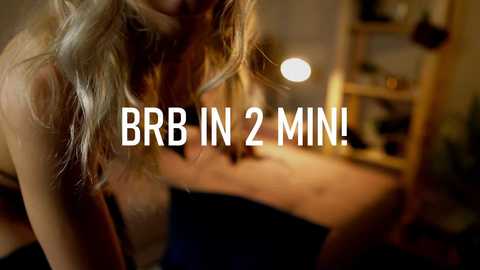 Video of a blonde woman in a dark room with blurred background, wearing a black top, and text overlay \"BRB IN 2 MIN!\