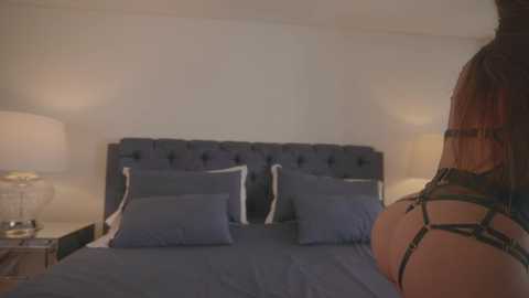 Media: A video of a woman in black lingerie standing next to a bed with blue bedding and white pillows. The room is dimly lit with a bedside lamp.