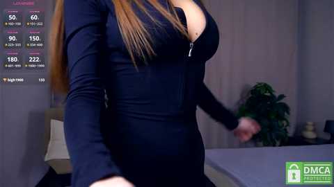 Video of a slim woman with long auburn hair in a tight, black zip-up dress, standing indoors near a desk and green plant.