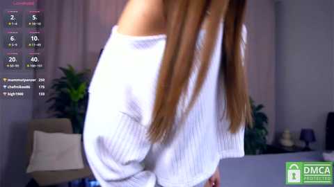 Video of a woman with long, straight brown hair, wearing a white, off-the-shoulder knit sweater and light-colored pants, standing in a modern, dimly lit living room.