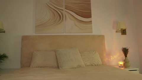 Media: Video of a minimalist bedroom with a beige upholstered headboard, two pillows, a decorative wall hanging, a side table with a candle, and a small potted plant. The room is warmly lit with a soft, ambient glow.