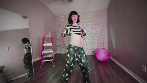 Video of a slender, pale-skinned woman with shoulder-length black hair, wearing a black and white striped crop top and camouflage pants, dancing energetically in a dimly lit, minimalist room with a pink exercise ball and a mirrored ladder.