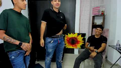 Media: Video of three young men in casual attire, one with a tattoo, standing in a room with a large yellow flower, pink wall, and a shelf with books and a pink poster.