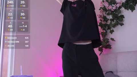 Media: Video of a person wearing a black crop top and high-waisted pants, holding a shirt against their chest, against a wall with green ivy and a pink floral arrangement.