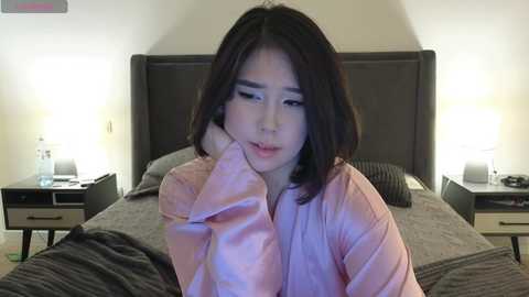 Media: Video of an Asian woman with shoulder-length dark hair, wearing a pink bathrobe, sitting on a bed with beige linens and two nightstands with lit lamps. She looks tired, with her head resting on her hand.