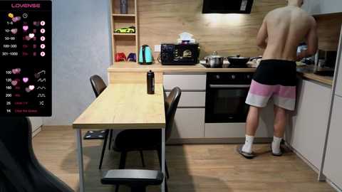 Media: A video of a young, shirtless man in black shorts standing on a wooden kitchen floor, facing a modern kitchen with light wood cabinetry and stainless steel appliances.