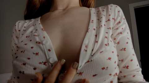 A video of a fair-skinned woman with long, auburn hair, wearing a low-cut, floral-patterned cardigan, exposing her cleavage. The background is dimly lit with a door visible.