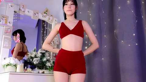 Media: Video of a slender, light-skinned Asian woman with short black hair, wearing a red bralette and high-waisted shorts, standing confidently in a bedroom with a mirror, floral arrangement, and fairy lights.