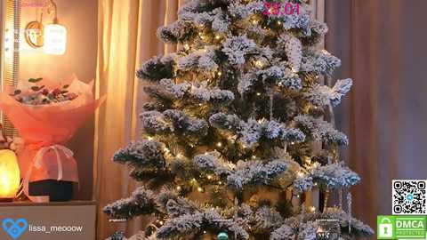 Media: Video of a snowy, artificial Christmas tree lit with warm, golden lights, set against a background of beige curtains and a lit candle in a glass holder.