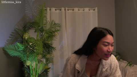 Media: A video of an Asian woman with long black hair, wearing a beige cardigan, standing next to a potted fern in a room with beige walls and white curtains.