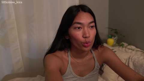 Media: Video of a young East Asian woman with long black hair, light skin, and a slim figure, wearing a grey tank top, blowing a kiss, in a cozy bedroom with a yellow plant.