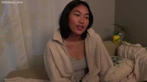 Media: Video of an Asian woman with medium-length black hair, wearing a beige robe over a gray tank top, sitting on a bed with floral bedding, in a dimly lit bedroom.