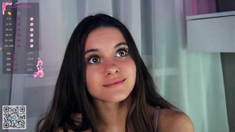 Media: Video of a young woman with long, dark brown hair, fair skin, and expressive eyes. She wears a light pink top, smiling slightly. Background includes a calendar with pink accents, a QR code, and soft, pastel-colored curtains.