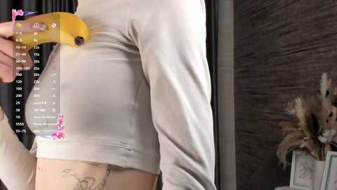 Media: Video of a person wearing a tight white long-sleeve shirt, holding a banana, with a chart overlay showing body temperature and weight. The background features dark wood paneling and a decorative floral arrangement.