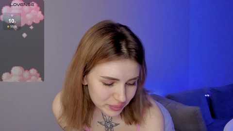 Media: Video of a young woman with shoulder-length, light brown hair, wearing a purple tank top, standing in a softly lit room with a blue accent. She has a tattoo on her neck and a poster with pink clouds in the background.