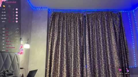 A video of a modern bedroom with beige textured curtains, a gray padded headboard, and blue LED strip lighting.