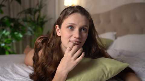 Media: Video of a fair-skinned, brunette woman lying on a bed, holding a green pillow with a thoughtful expression, in a softly lit, cozy bedroom.