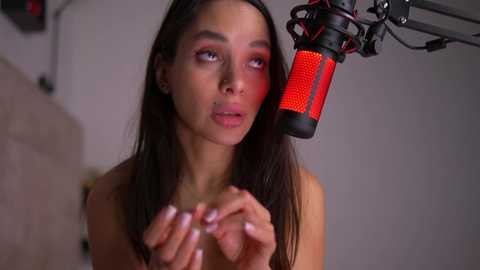 Media: Video of a topless, light-skinned woman with long dark hair and red makeup, holding hands in front of a red microphone in a studio.