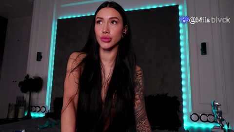 Video of a light-skinned woman with long black hair, wearing a black sheer top, standing indoors with a tattooed arm, surrounded by neon blue lights, in a modern room.