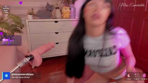 Video of a young woman with long black hair, wearing a tight gray crop top and pink skirt, kneeling on a shiny wooden floor. She has a shocked expression as a large, realistic-looking dildo is thrust towards her face.