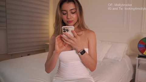 Video of a young woman with light skin and long, straight brown hair, wearing a white strapless dress, sitting on a bed, using a white iPhone.