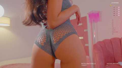 Media: Video of a woman with long brown hair, wearing a teal, fishnet bodysuit, showcasing her toned buttocks. Background features pink walls, a pink chair, and a pink lamp.