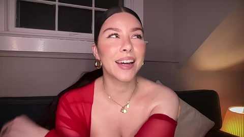Media: Video of a smiling, fair-skinned woman with dark hair, wearing a red off-shoulder dress, gold necklace, and earrings, sitting indoors near a window and lamp.