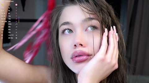 Media: Video of a young woman with wet, long brown hair and striking green eyes, wearing glossy pink lipstick, looking introspective. She rests her hand on her face, set against a blurred, modern background with pink and red accents.