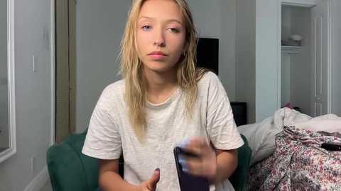 Media: Video of a young blonde girl in a white T-shirt, holding a smartphone, in a messy bedroom with a green couch and floral bedspread.