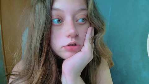 Media: Video of a young, fair-skinned girl with long, wavy brown hair, blue eyes, and a contemplative expression, resting her chin on her hand against a blurred teal background.