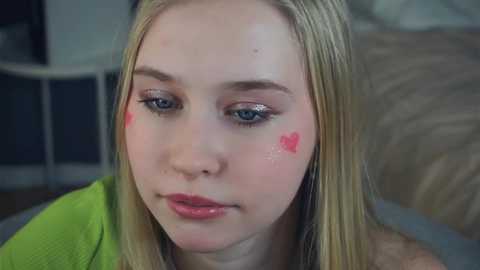 Media: Video of a young Caucasian girl with light skin, blonde hair, blue eyes, wearing a green top. She has pink heart-shaped face paint on her cheek. Background shows a blurry, indoor setting with beige furniture.