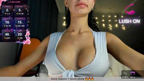 Media: Video of a light-skinned woman with long black hair, wearing a low-cut, light blue halter top, revealing ample cleavage. Background features a dark room with a glowing \"Lush On\" sign and a digital weather display.