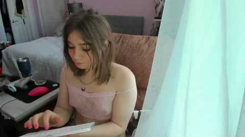 Video of a young, fair-skinned woman with shoulder-length brown hair, wearing a pale pink off-shoulder top, sitting on a couch, playing with a remote, in a cluttered living room.