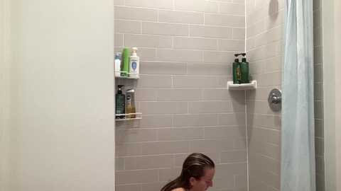 Media: A video of a modern bathroom with beige subway tiles, green shampoo bottles on a shelf, a showerhead, and a person with wet hair partially visible in the bottom left corner.